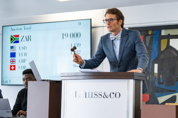 Auctioneer in a blue blazer and bow tie at a podium, presenting during an auction with a large screen showing totals behind him.